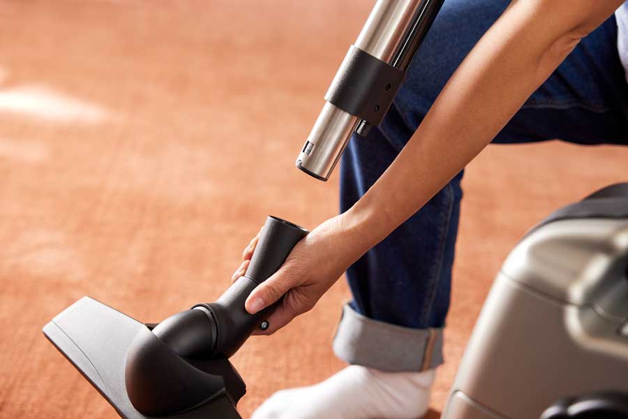 A close-up of a person attaching a black floor nozzle to a hose of a Kirby Signature Series C550 canister vacuum. The nozzle is positioned over a terracotta-colored carpet.