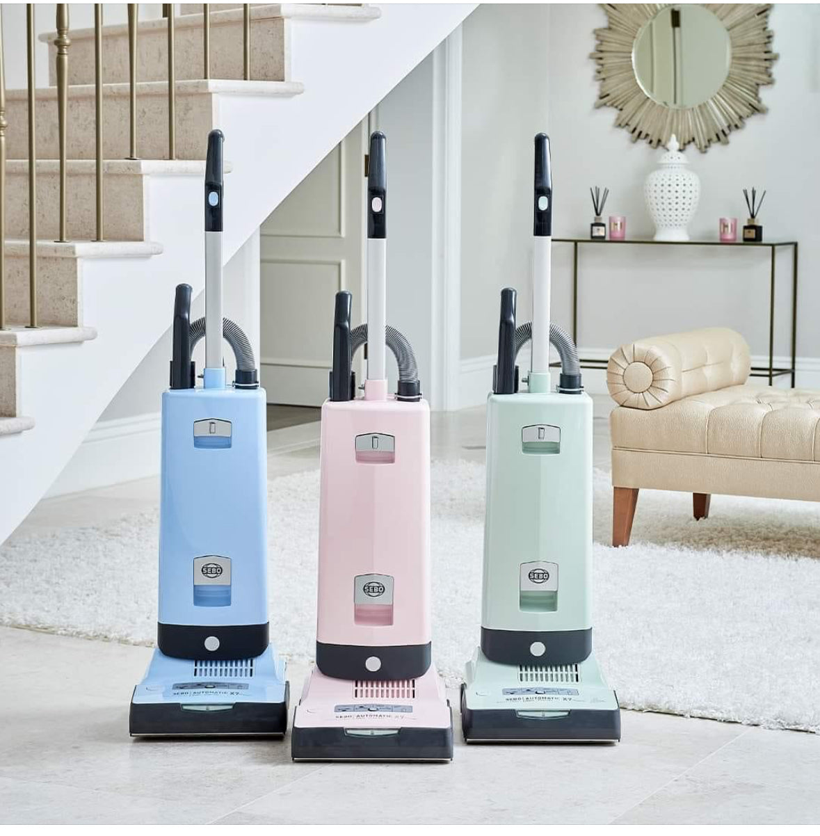 Three vacuum cleaners in pastel colors on a carpeted floor with a staircase and living room in the background.