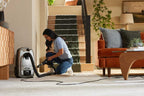 A woman kneels on a light beige carpet in a modern living room, using a Kirby Signature Series C530 canister vacuum cleaner with its hose connected. The room features a staircase with green carpeting and a orange and brown sofa.