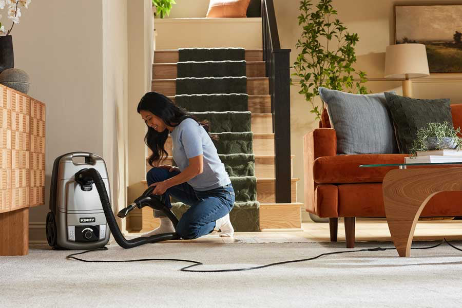 A woman kneels on a light beige carpet in a modern living room, using a Kirby Signature Series C530 canister vacuum cleaner with its hose connected. The room features a staircase with green carpeting and a orange and brown sofa.