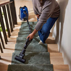 A woman using a black Kirby Signature Series C550 canister vacuum to clean wooden stairs with green carpet runners.