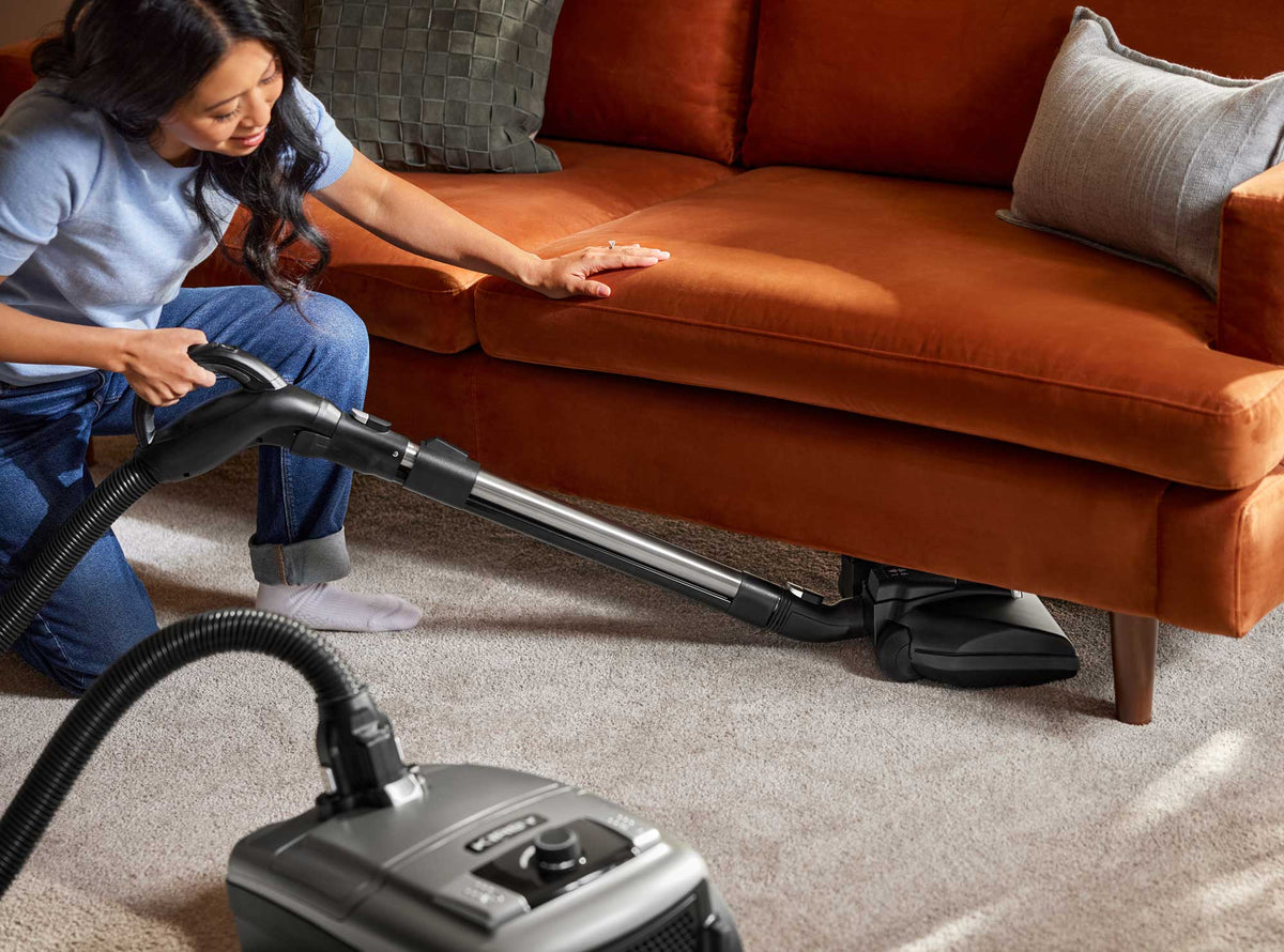 A woman kneels beside an orange sofa in a living room, using a Kirby Signature Series C550 canister vacuum cleaner to clean the carpet and sofa base.
