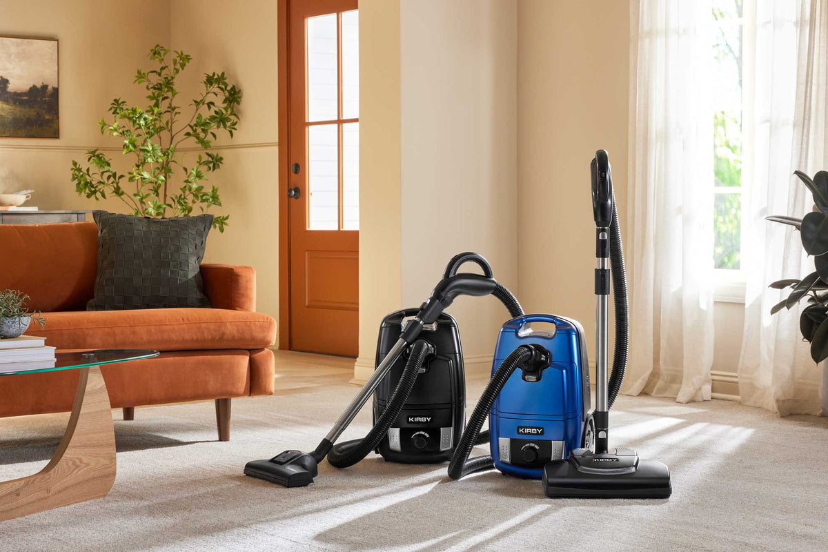Kirby Signature Series C550 and C530 canister vacuums, one black and one blue, displayed side-by-side on a beige carpet in a bright, modern living room.