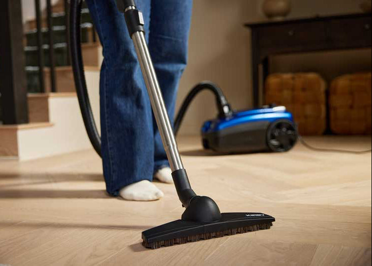 A person wearing blue jeans and white socks uses a Kirby Signature Series C550 canister vacuum cleaner with a black wand and floor head to clean a light wood herringbone floor in a home interior setting.