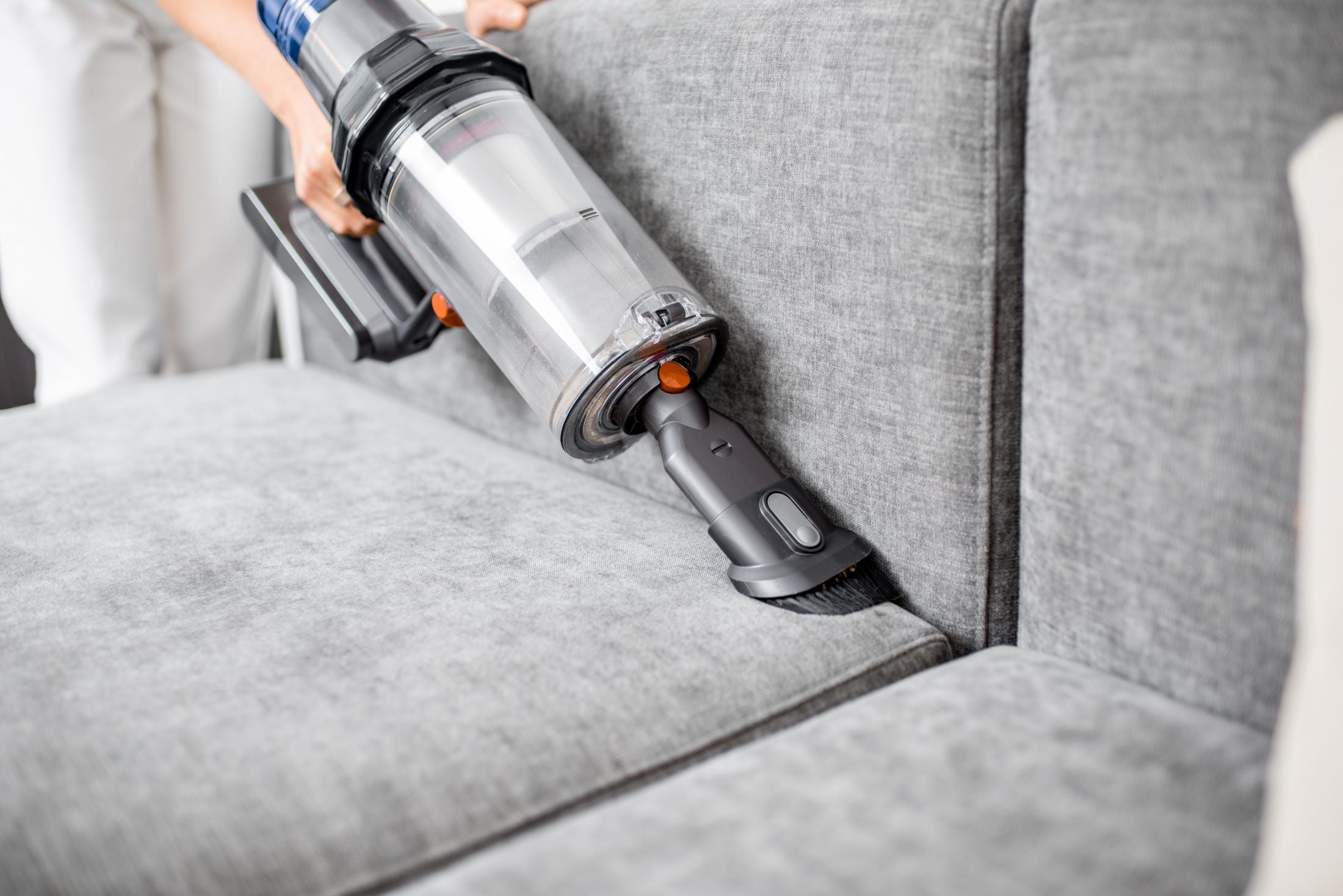 Person using a handheld vacuum cleaner to clean a gray sofa.