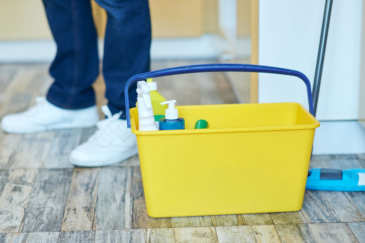 Yellow cleaning bucket with supplies on a wooden floor, person in blue pants and white shoes partially visible.