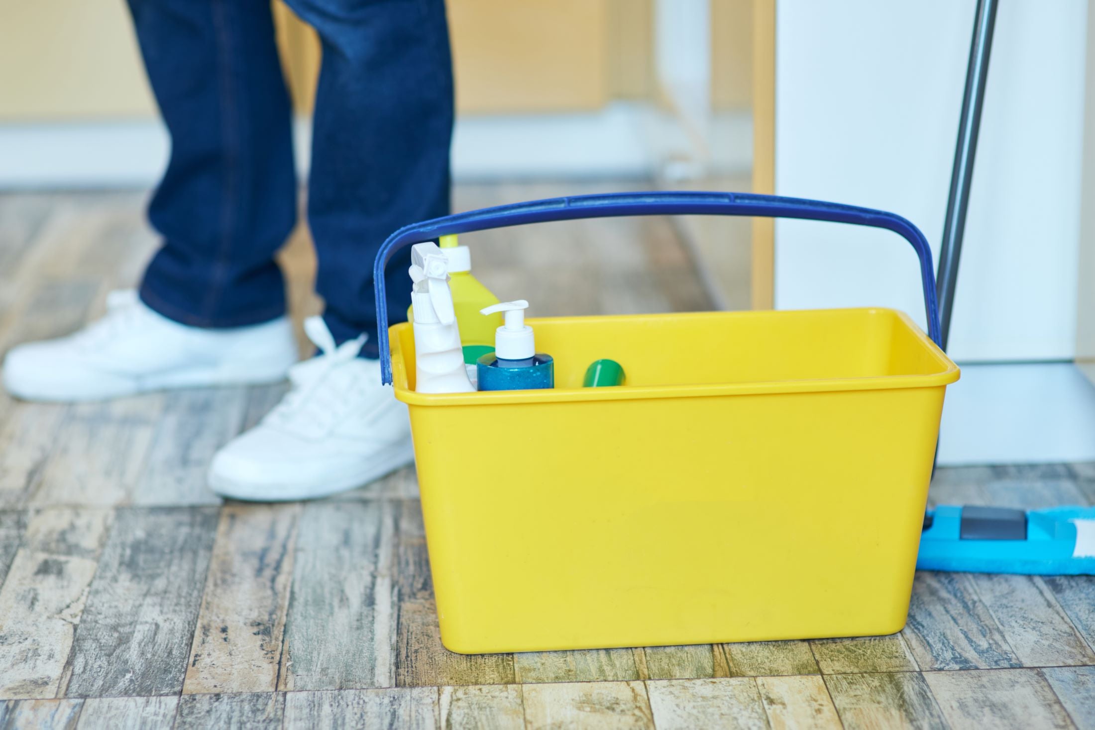 Yellow cleaning bucket with supplies on a wooden floor, person in blue pants and white shoes partially visible.