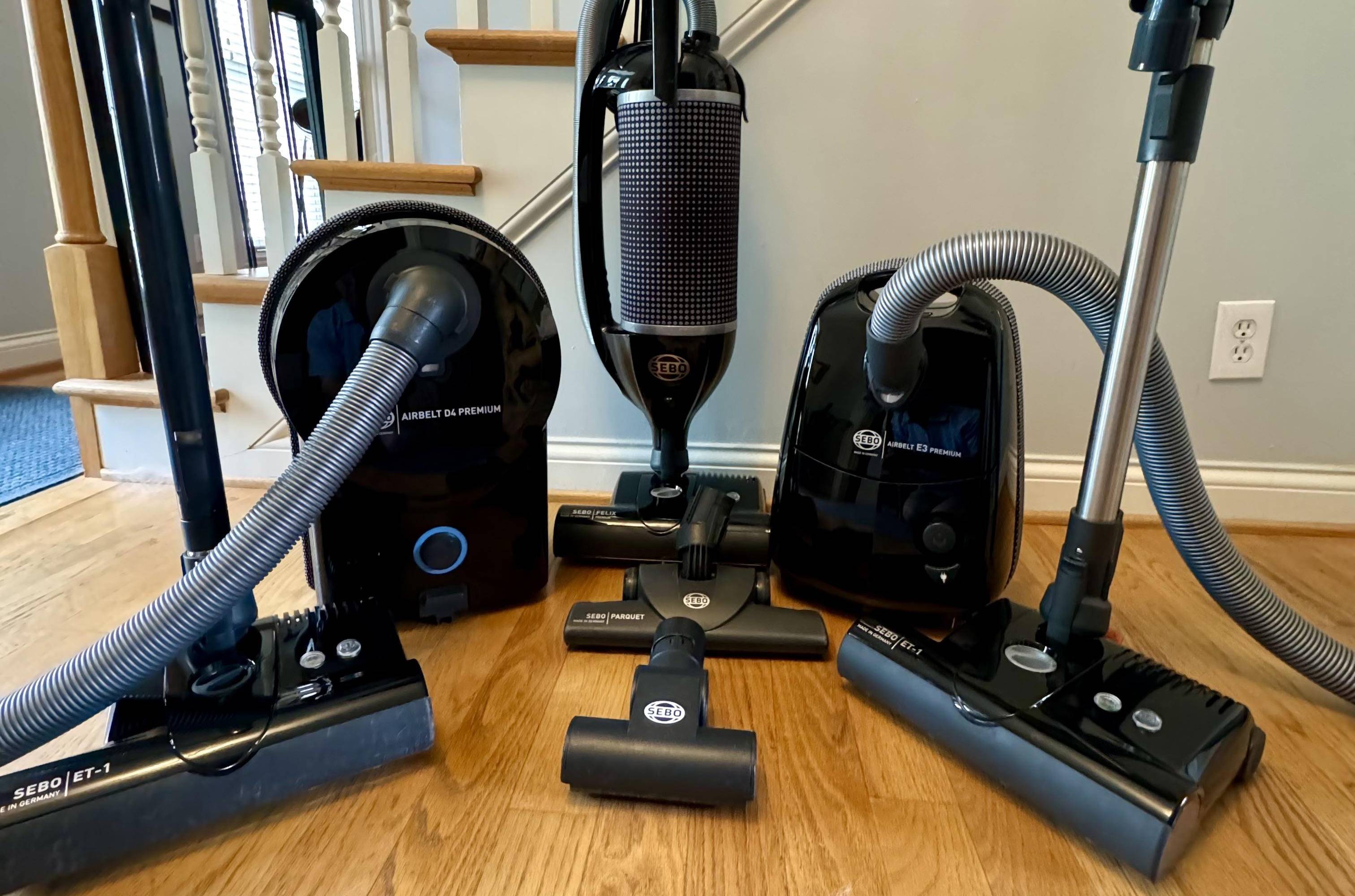 Four different vacuum cleaner models on a wooden floor with a staircase in the background.